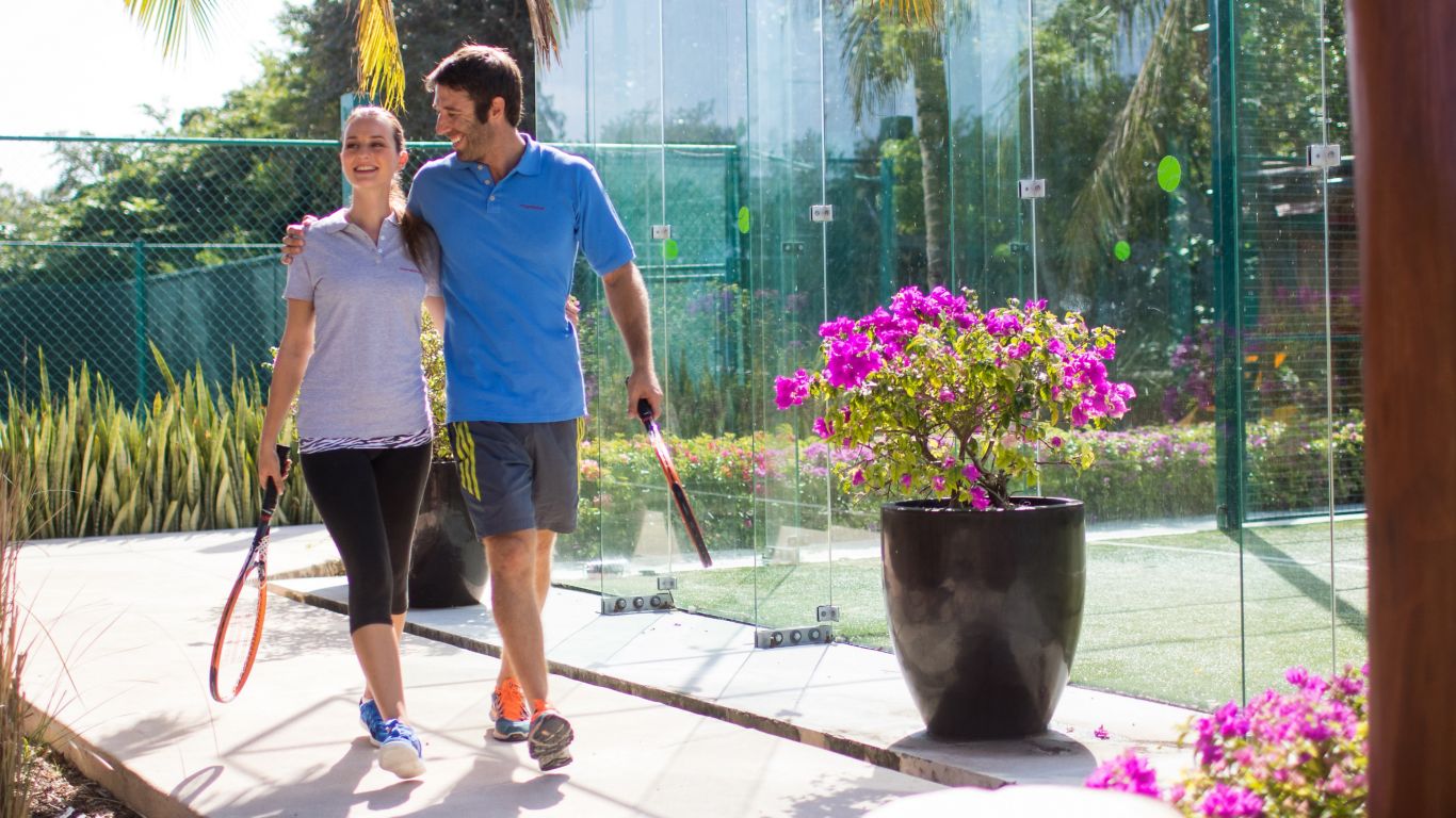 woman and man holding tennis racquets walking with their arms around one in front of tennis courts in athletic clothing  